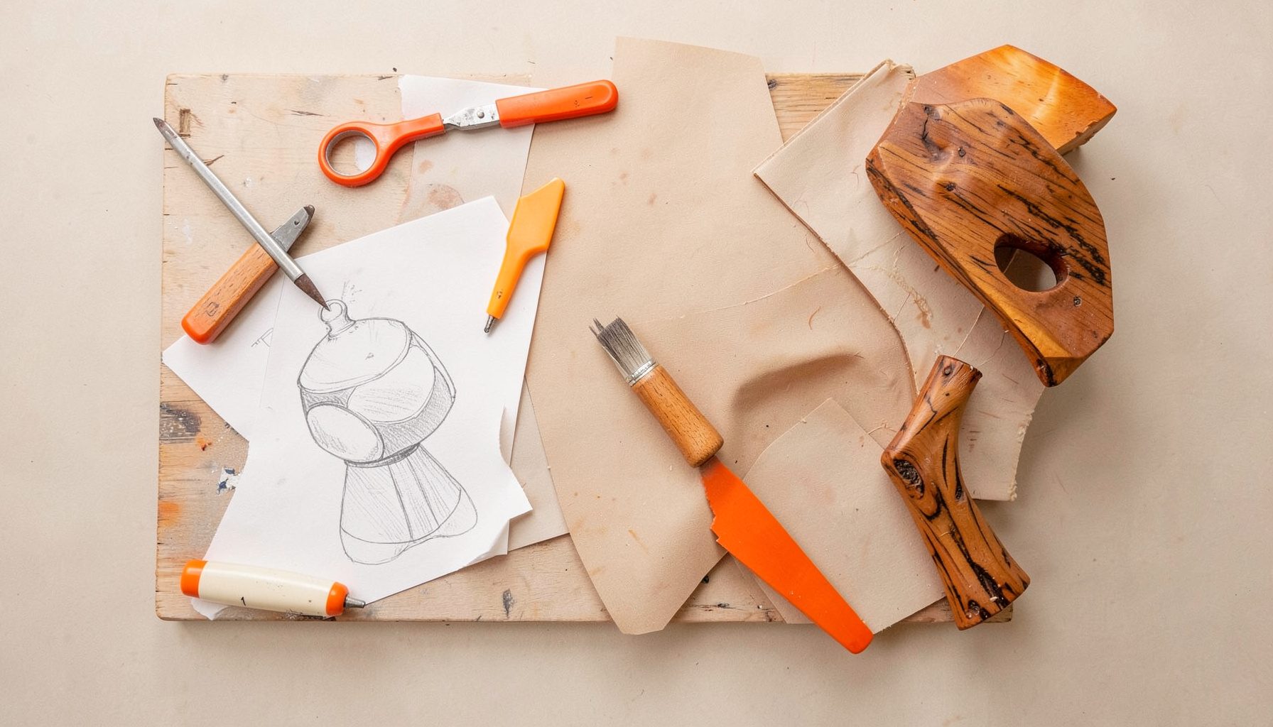 Overhead flat-lay photograph of an artisan workbench with tools, sketches and a half-finished sculptural piece in warm wood tones, soft natural light